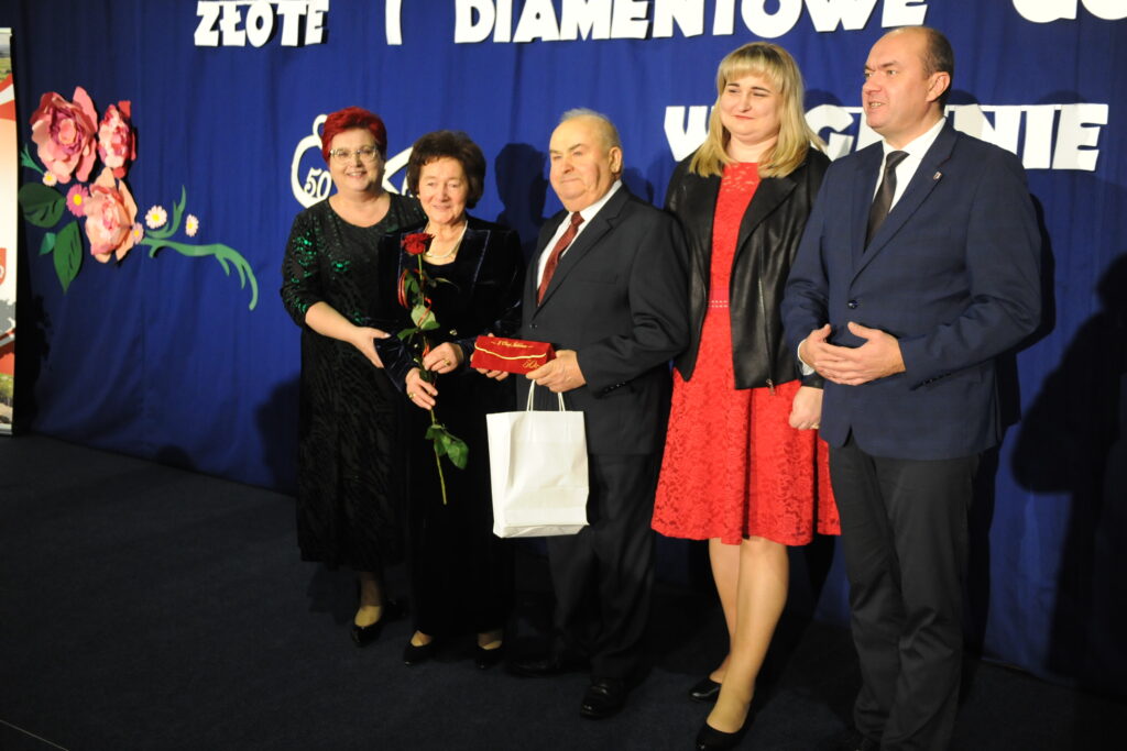 Pięć osób stoi na tle niebieskiej kurtyny z dekoracjami kwiatowymi. Dwie z nich trzymają różę i biały prezent. Cztery osoby są elegancko ubrane, jedna w czerveną sukienkę.