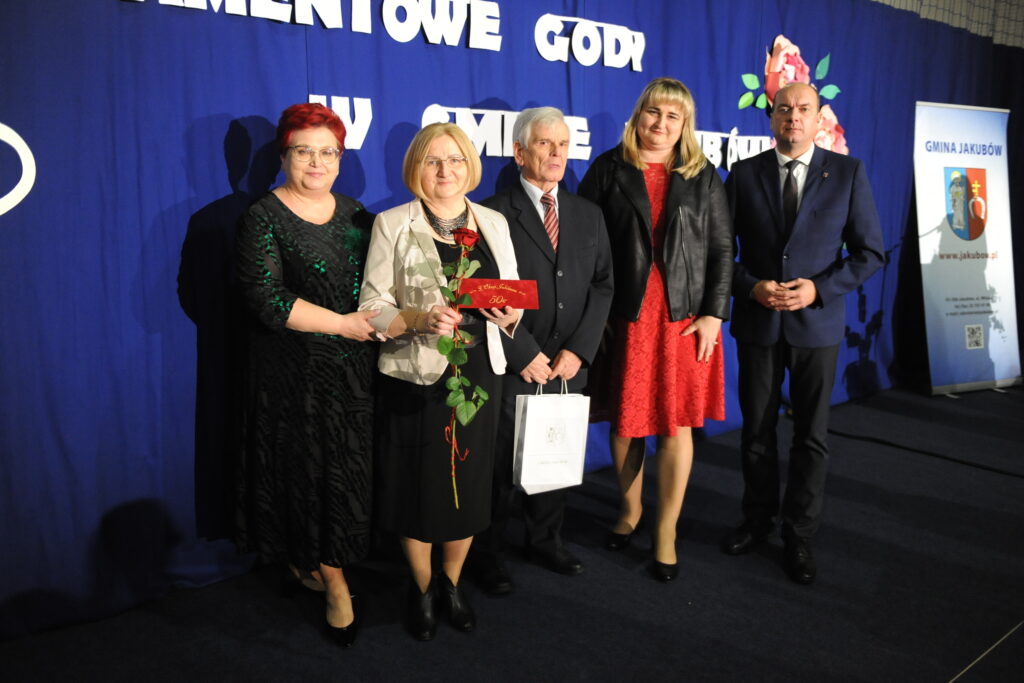 Na niebieskim tle stoi pięć osób w eleganckich strojach. Kobieta trzyma czerwoną różę i kopertę. Obok jest baner z napisem Gmina Jakubów.