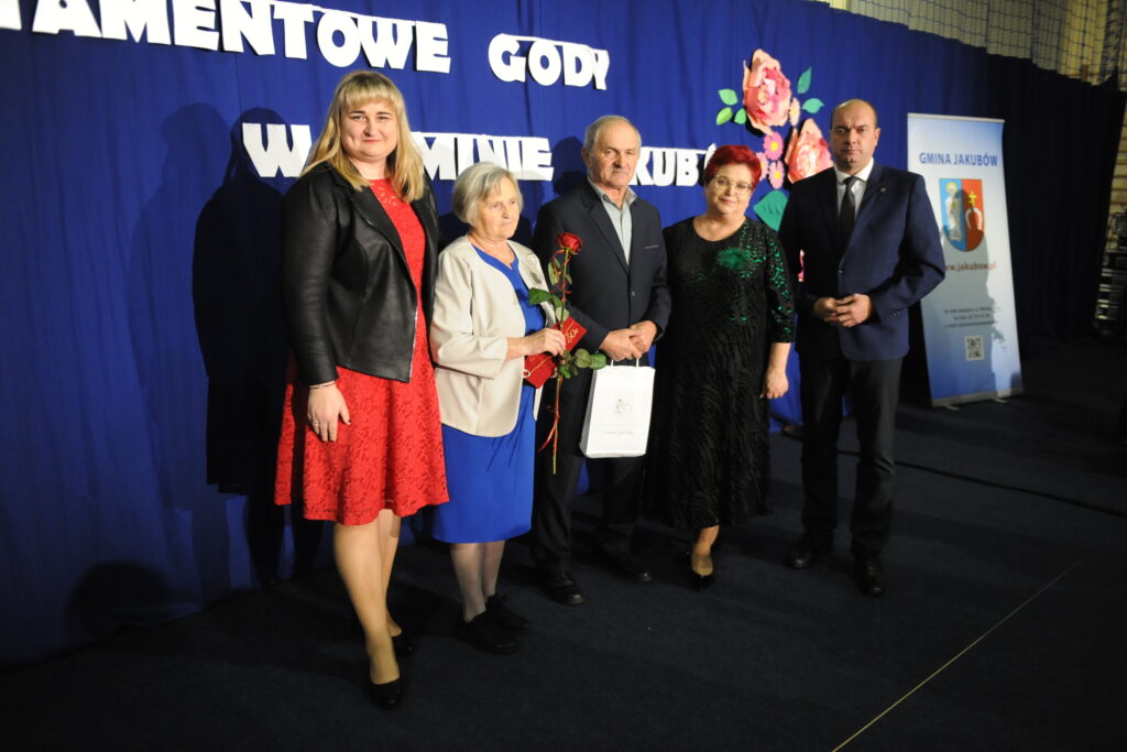 Pięć osób stoi na tle niebieskiej zasłony z napisem, kobieta trzyma różę. W tle kwiaty i plakat z informacją.