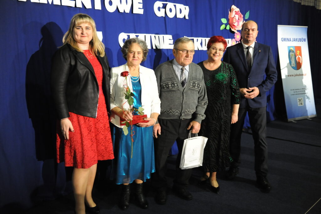 Pięć osób pozuje na tle niebieskiej kurtyny z napisem. Są odświętnie ubrani i trzymają róże i torby. Po prawej stoi baner z napisem Gmina Jakubów.