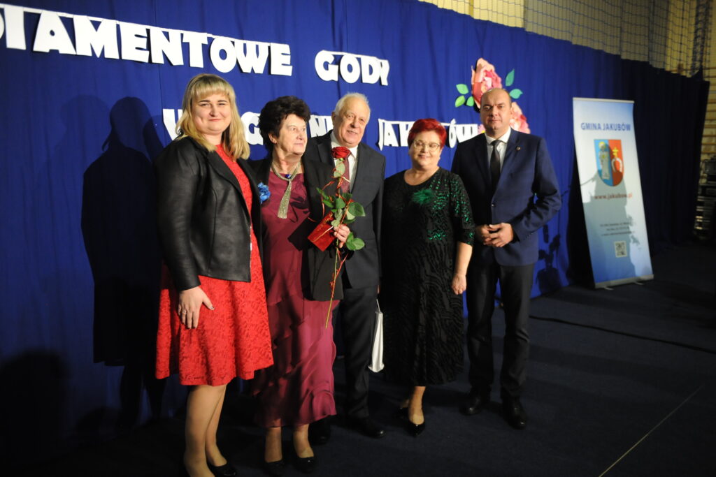 Pięć osób pozuje na tle niebieskiej zasłony z napisem. Kobieta trzyma różę, obok banner gminy. Eleganckie stroje, uroczystość.