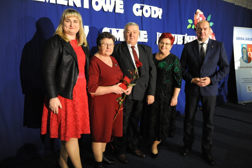 Grupa pięciu osób pozuje na tle niebieskiej kurtyny. Kobieta trzyma czerwoną różę. W tle napis i logo gminy.