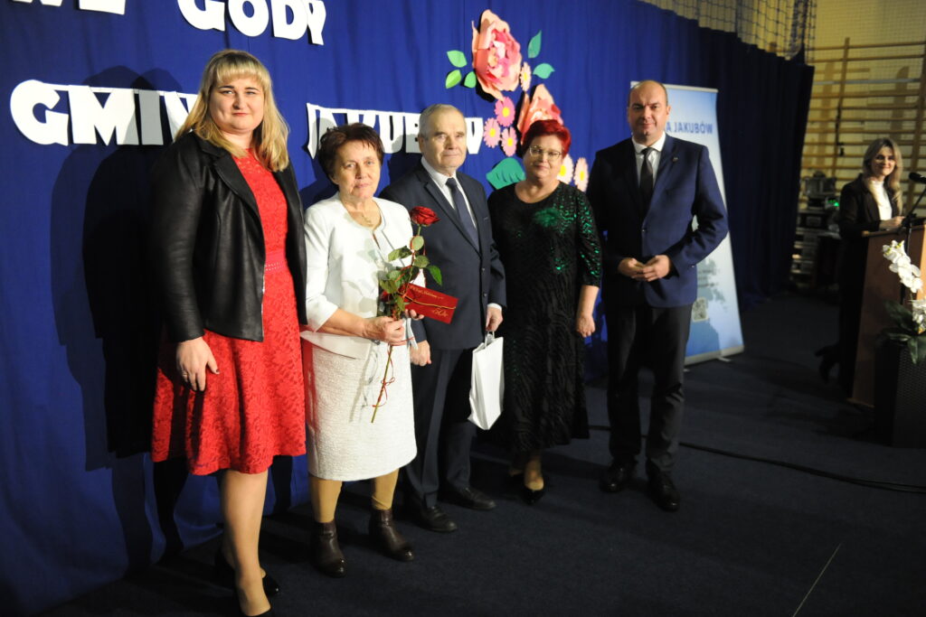 Pięć osób stoi na tle niebieskiej ściany z kwiatami. Wszyscy elegancko ubrani, jedna z kobiet trzyma czerwoną różę. Obok nich widoczny jest stojak z napisem.