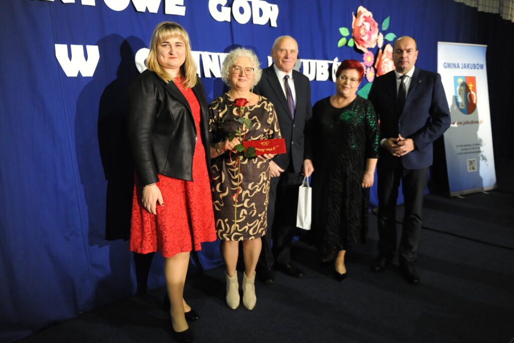 Pięć osób stoi na tle niebieskiej zasłony z białym napisem. Kobieta w środku trzyma różę i czerwoną kopertę. Po prawej stronie widoczny jest plakat z napisem Gmina Jakubów.