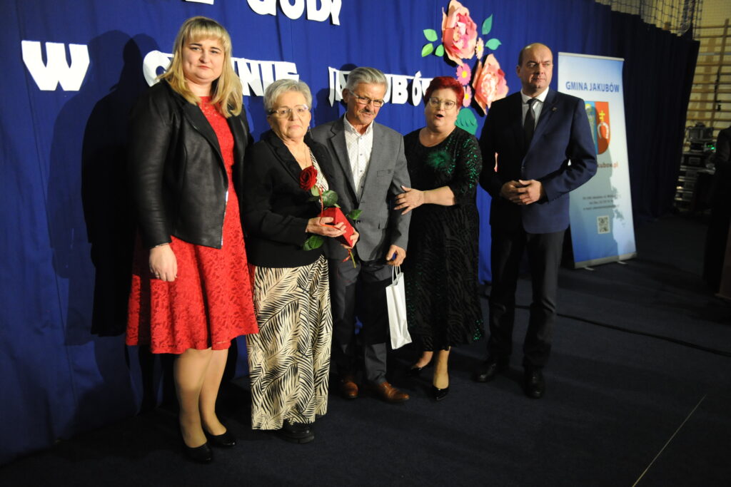 Grupa pięciu osób pozuje na tle granatowej kurtyny z kwiatami. Starsza kobieta trzyma czerwoną różę. Obok banner Gminy Jakubów.