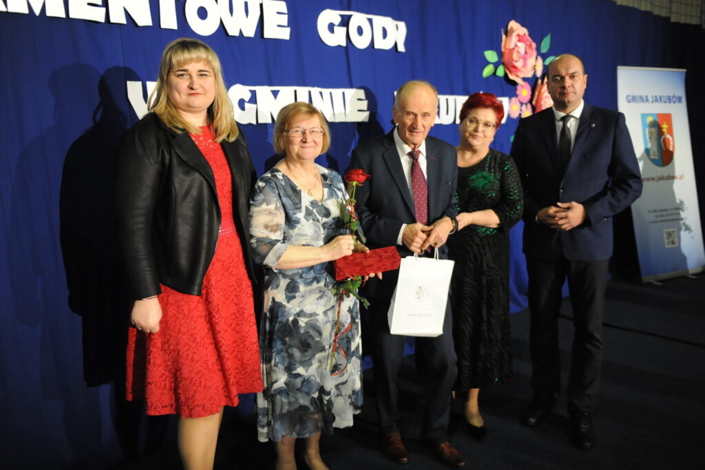 Na zdjęciu jest pięć osób na tle niebieskiej ściany z napisami. Kobieta w czerwonej sukni trzyma białą torbę. Obok stoi starsza para z różą.