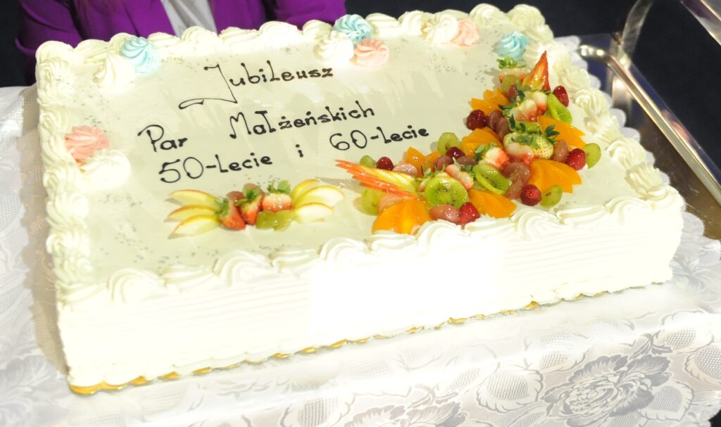 Prostokątny tort z napisem Jubileusz 50-lecie i 60-lecie. Dekoracje z owoców: kiwi, truskawki, winogrona, pomarańcze. Tło białe, imituje obrus.