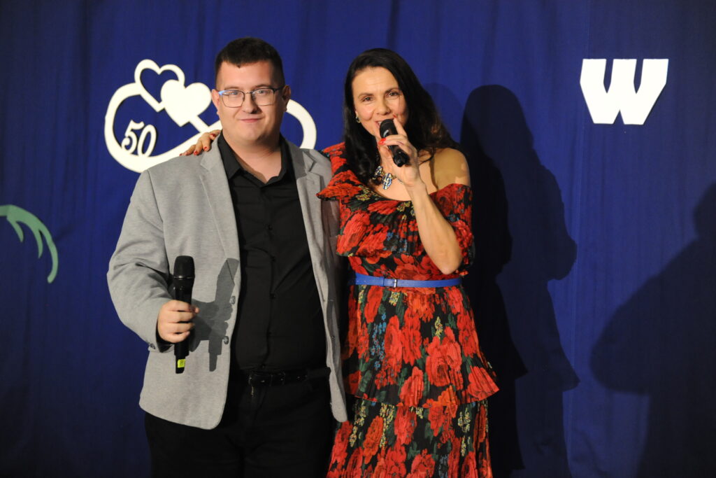 Dwie osoby z mikrofonami, mężczyzna w szarej marynarce i kobieta w czerwonej sukni. Niebieskie tło z białymi symbolami. Ujęcie z wydarzenia.