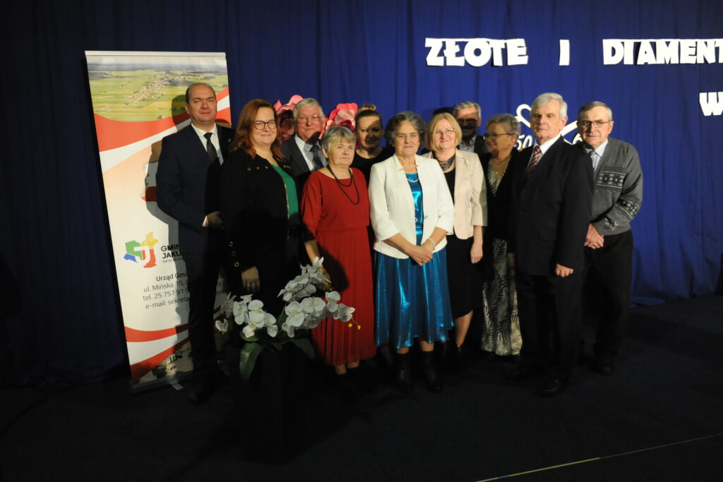 Grupa osób na tle niebieskiej scenografii, z napisem Złote i Diamenty. Ubrani elegancko, stoją obok dużego plakatu. Przed nimi biała orchidea w doniczce.