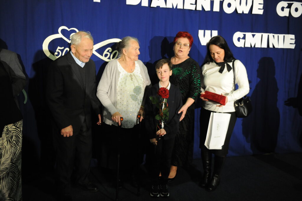 Grupa ludzi pozuje na tle niebieskiej dekoracji z białymi napisami. Starsze osoby wspiera chłopiec trzymający różę. Jeden z dorosłych trzyma torbę upominkową.