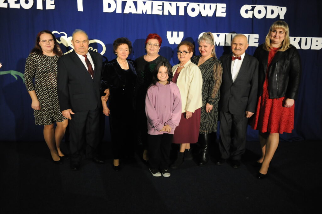 Grupa osób pozuje na tle niebieskiej ściany z białym napisem; są elegancko ubrani. Na pierwszym planie stoi dziecko w fioletowej bluzie. Atmosfera jest uroczysta.