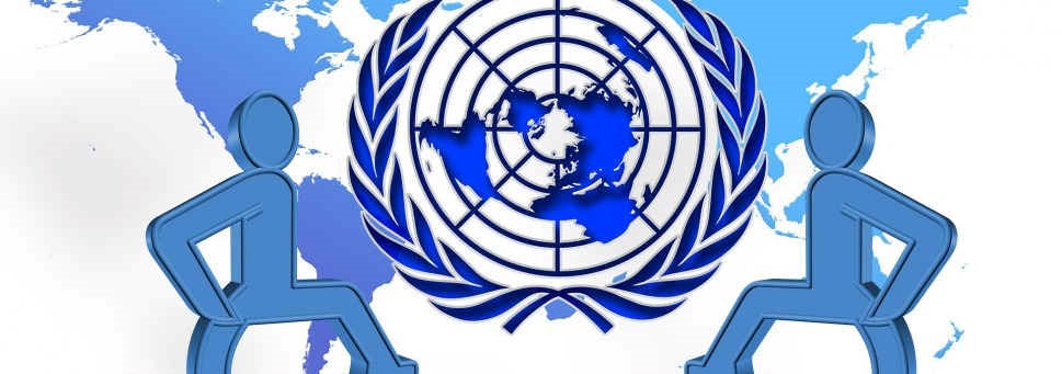 Mapa świata w tle, na pierwszym planie logo ONZ w kolorze niebieskim. Po bokach dwie niebieskie sylwetki. Kolorystyka zdominowana przez odcienie niebieskiego i biel.