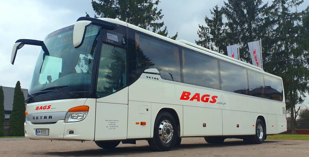 Na zdjęciu znajduje się biały autobus z czerwonym napisem BAGS. Stoi na tle drzew i pochmurnego nieba. Przód autobusu ma pomarańczowe reflektory.
