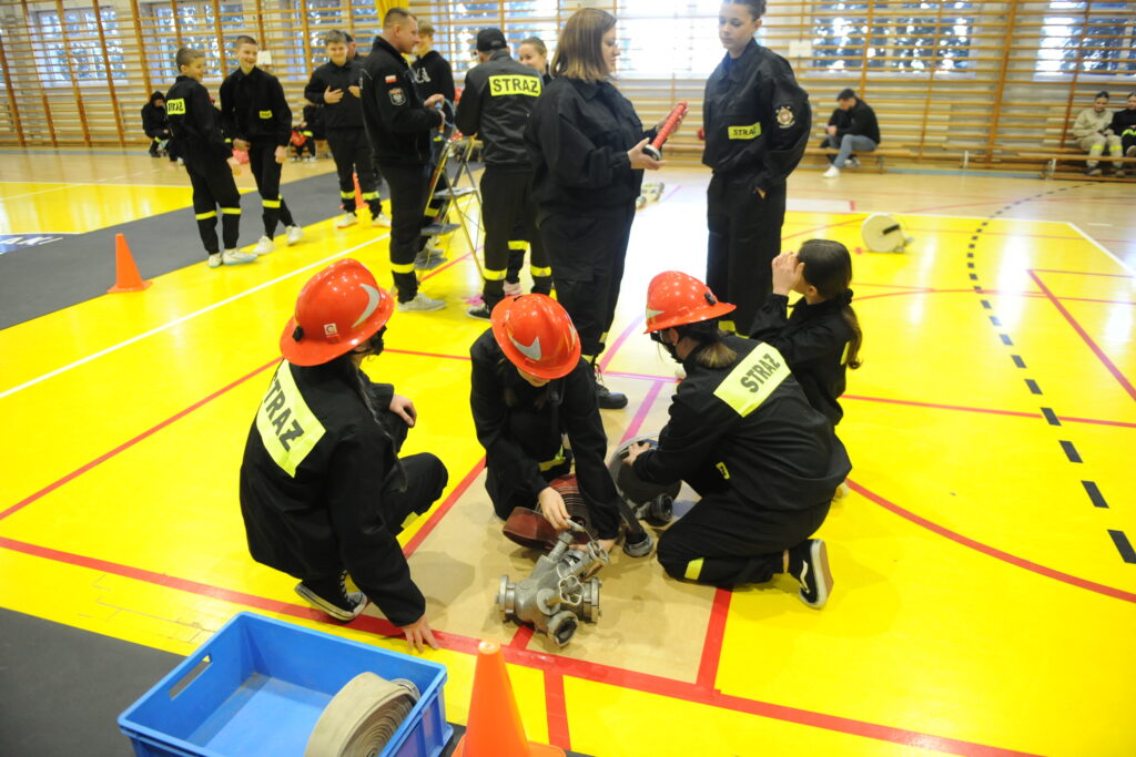 Grupa młodych ludzi w strojach strażackich ćwiczy na żółtej sali gimnastycznej. Noszą czerwone kaski i używają sprzętu. W tle widać drabinki gimnastyczne.