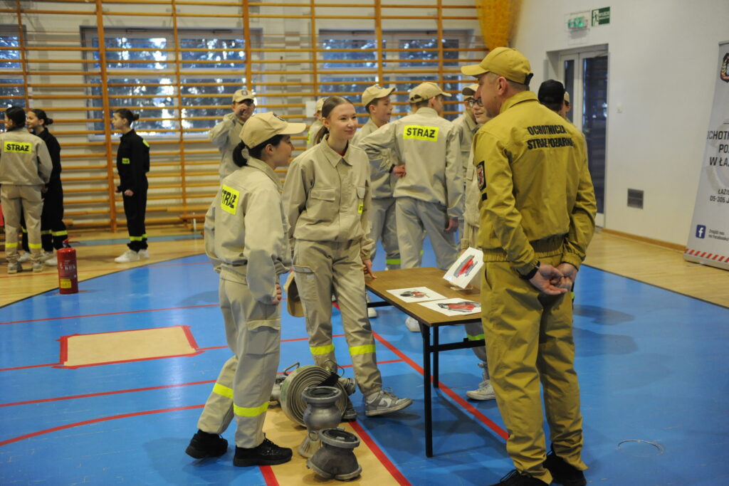Grupa młodych ludzi i strażak w mundurach stoją na niebieskiej podłodze sali gimnastycznej, rozmawiając przy stole z dokumentami.