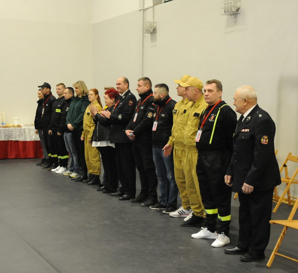 Grupa osób stoi w szeregu, ubrani w uniformy w odcieniach czerni i żółtego. Tło jasne z białą ścianą, obok stół z białym obrusem. Osoby mają czerwone identyfikatory.