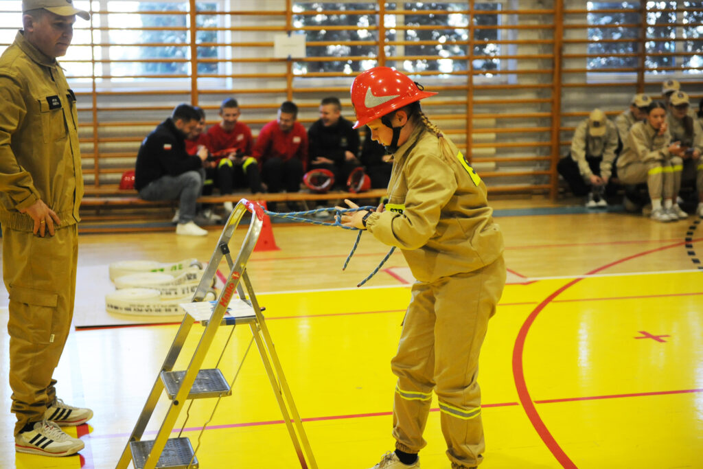 Dziewczynka w czerwonym hełmie ćwiczy wiązanie liny przy drabince. Ma na sobie beżowy strój strażaka. W tle widać obserwujących ludzi i salę gimnastyczną.