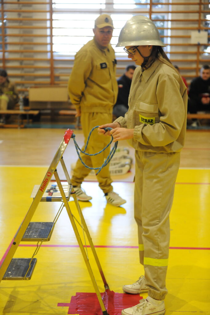 Osoba w stroju strażaka stoi przy drabince, trzymając linę. Ma na sobie hełm, na żółtym tle sali gimnastycznej. W tle widać innych uczestników.