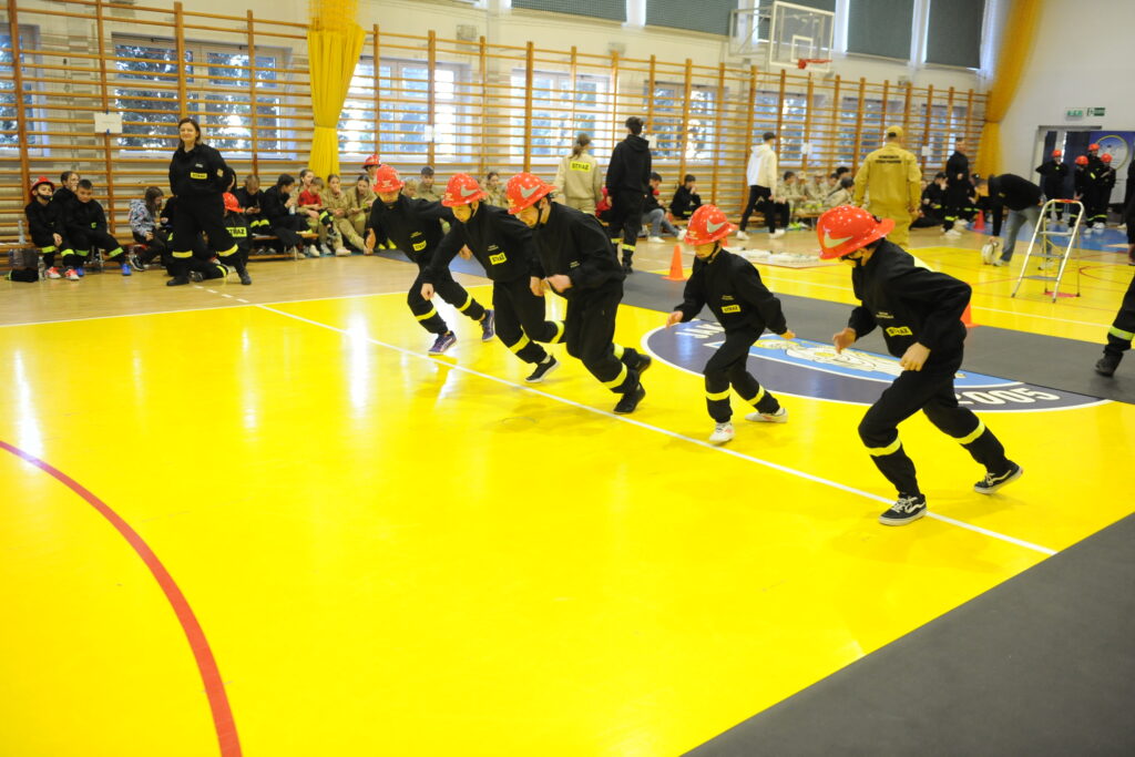 Na zdjęciu grupa dzieci w hełmach strażackich biegnie po żółtej podłodze sali gimnastycznej. W tle widać siedzących ludzi w ciemnych ubraniach. Ściany są wyłożone drewnianymi drabinkami.