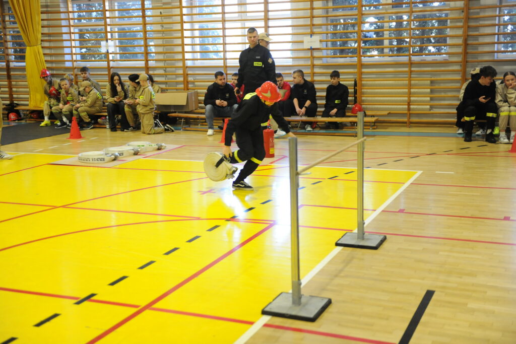 Na zdjęciu widać dzieci w strojach strażackich w sali gimnastycznej. Tło w tonacji jasnej, podłoga ma żółte pola. Dzieci ćwiczą zwinność.