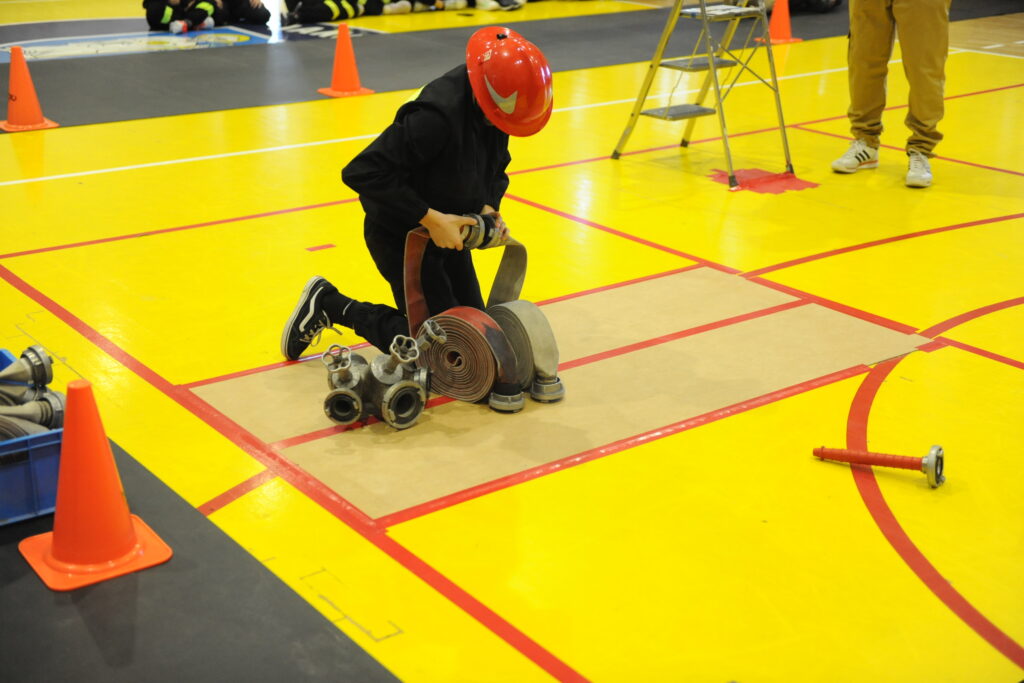 Dziecko w czerwonym kasku klęczy na żółtej podłodze, układając węże strażackie. Otoczenie oznakowane czerwonymi liniami. Widoczna lekka drabinka i pachołki.