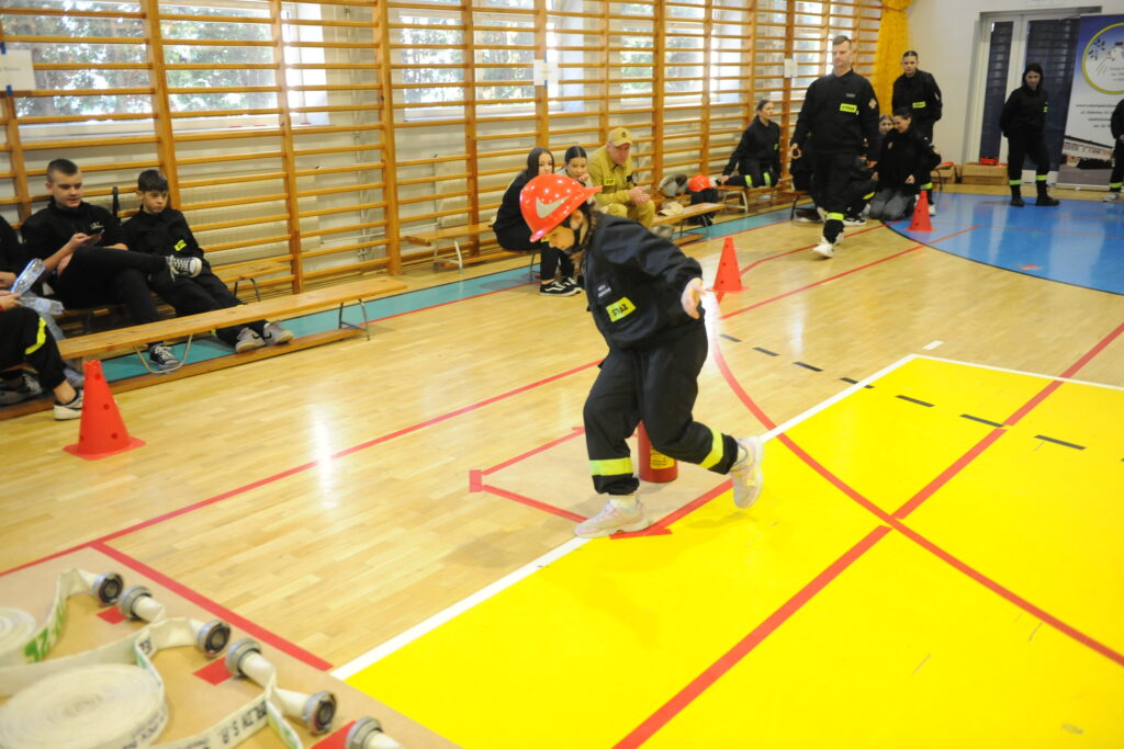 Dziecko w stroju strażaka biegnie z wężem na sali gimnastycznej. Podłoga jest drewniana z żółtymi i czerwonymi liniami. W tle siedzą ludzie przy ścianie z drabinkami.