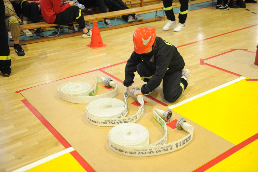 Osoba w czerwonym hełmie układa wąż strażacki na sali gimnastycznej. Podłoga jasna, w tle ławka i widownia. Atmosfera ćwiczeń.