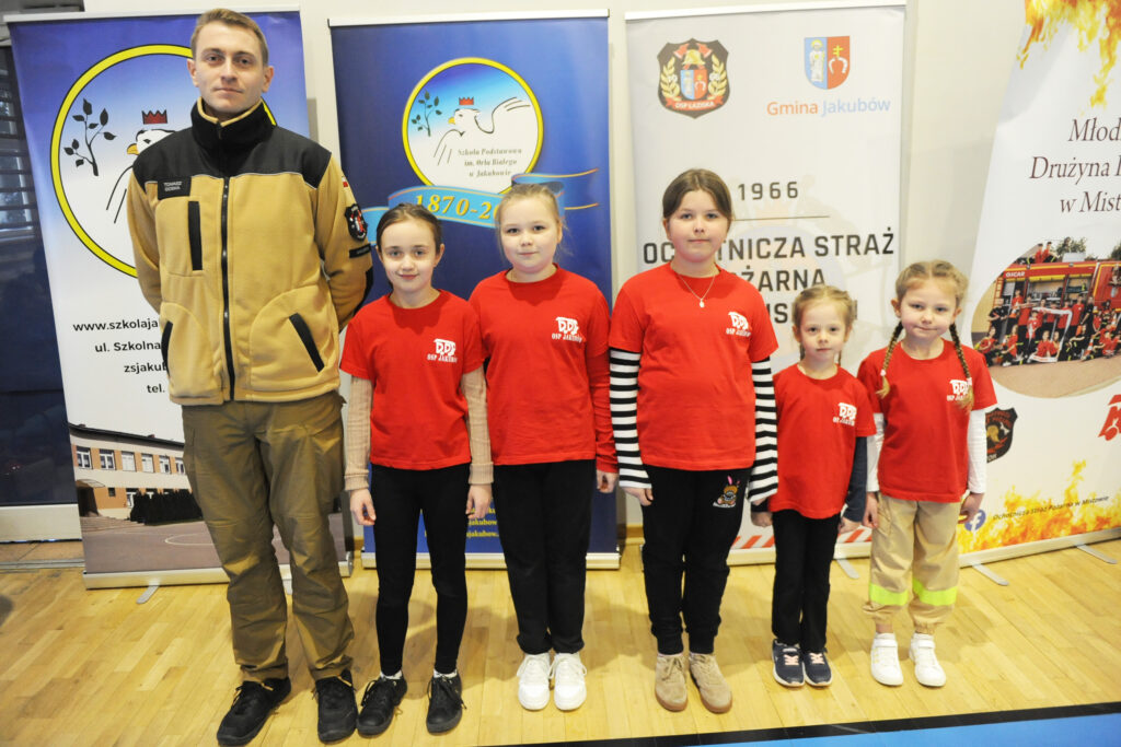 Grupa osób stoi na drewnianej podłodze przed kolorowymi bannerami. Mają na sobie czerwone koszulki. Tło jest jasne z różnymi grafikami i napisami.