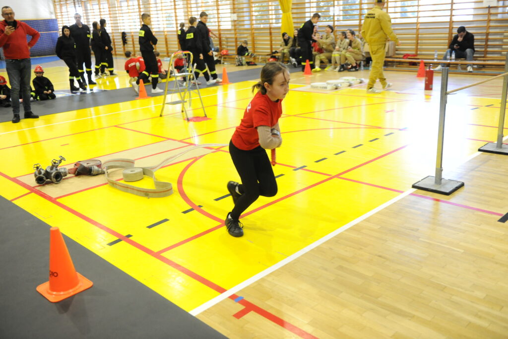Dziewczynka w czerwonej koszulce biegnie w hali sportowej z żółtym parkietem. W tle strażacy w czarnych i żółtych strojach. Obok niej stoją sprzęty strażackie.