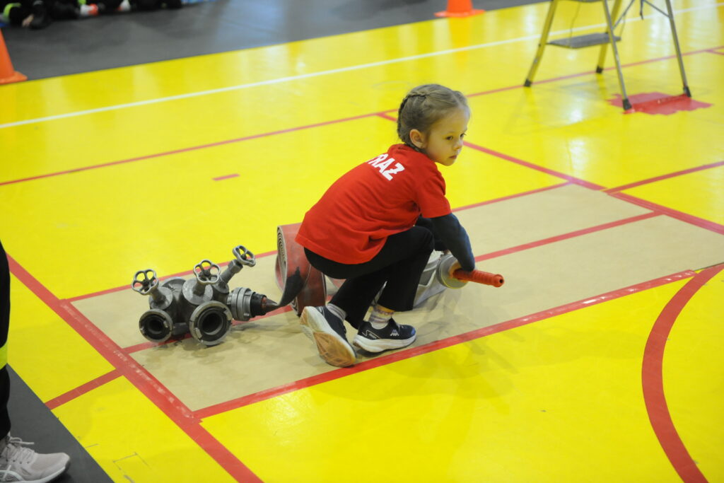 Dziewczynka w czerwonej koszulce siedzi na żółtej podłodze. Trzyma wąż strażacki obok zaworów. W tle są pomarańczowe pachołki.
