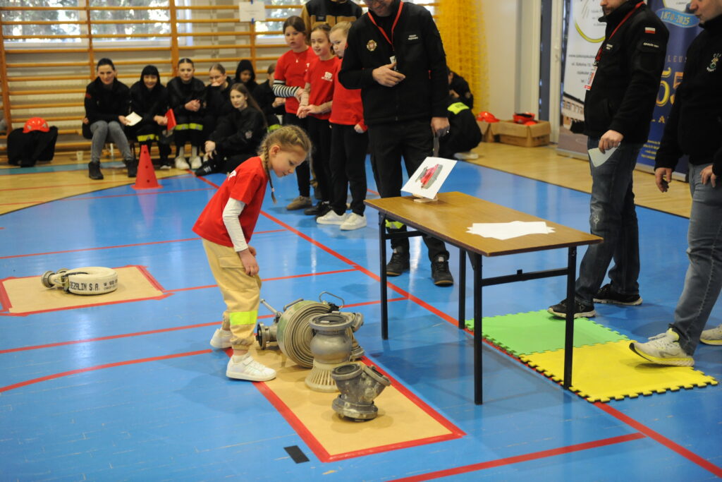 Dziewczynka w czerwonej koszulce stoi obok sprzętu strażackiego na niebieskiej podłodze. W tle grupka dzieci i dorośli. W sali dominują kolory czerwony, niebieski i żółty.