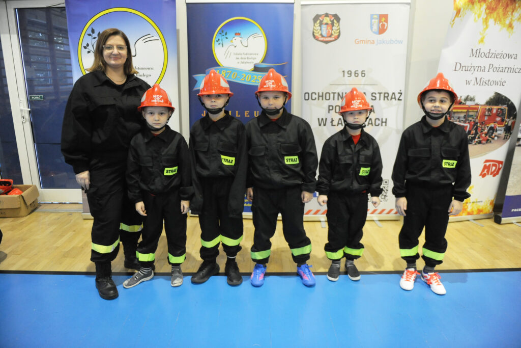 Grupa dzieci w strażackich mundurach i kaskach stoi obok kobiety. Tło stanowią niebieskie i białe bannery. Podłoga jest jasna, drewniana.