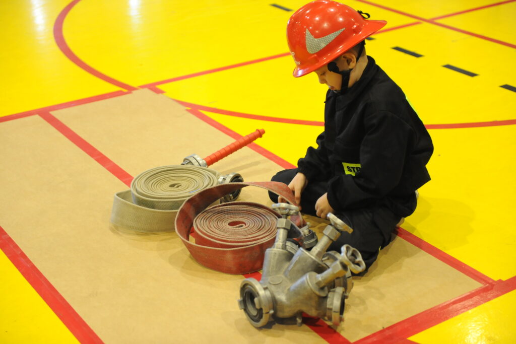 Dziecko w czerwonym hełmie strażackim siedzi na żółtej macie. Przed nim leżą zwinięte węże strażackie i rozdzielacz. Tło to wnętrze sali z czerwonymi liniami.