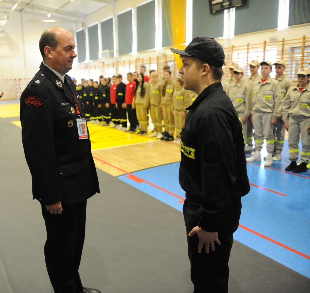 Na obrazie widać uroczysty apel w hali sportowej. Mężczyzna w mundurze strażaka stoi naprzeciw młodego chłopaka. Tło to rząd osób w różnokolorowych uniformach na parkiecie.