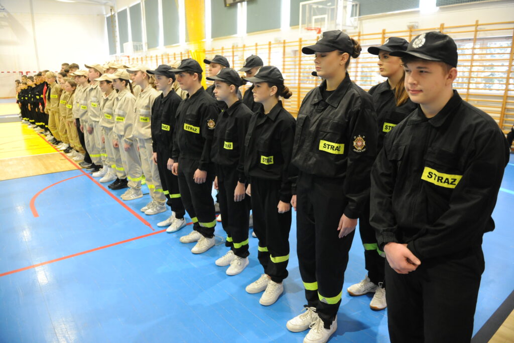 Grupa młodych ludzi w mundurach strażackich stoi w szeregu na sali gimnastycznej. Stroje są czarne i beżowe z napisem STRAŻ. Tło to niebieska podłoga i jasne ściany z oknami.