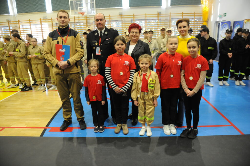 Grupa dzieci i dorosłych na sali gimnastycznej. Dzieci w czerwonych koszulkach, obok przedstawiciele straży pożarnej. Tło z ludźmi w mundurach strażackich.