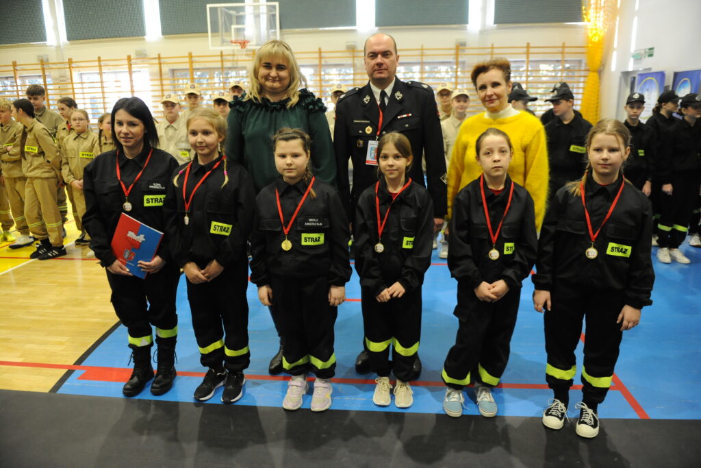 Grupa dzieci w czarnych mundurach straży z medalami, na tle szkolnej hali sportowej, obok dorosłych w strojach cywilnych.