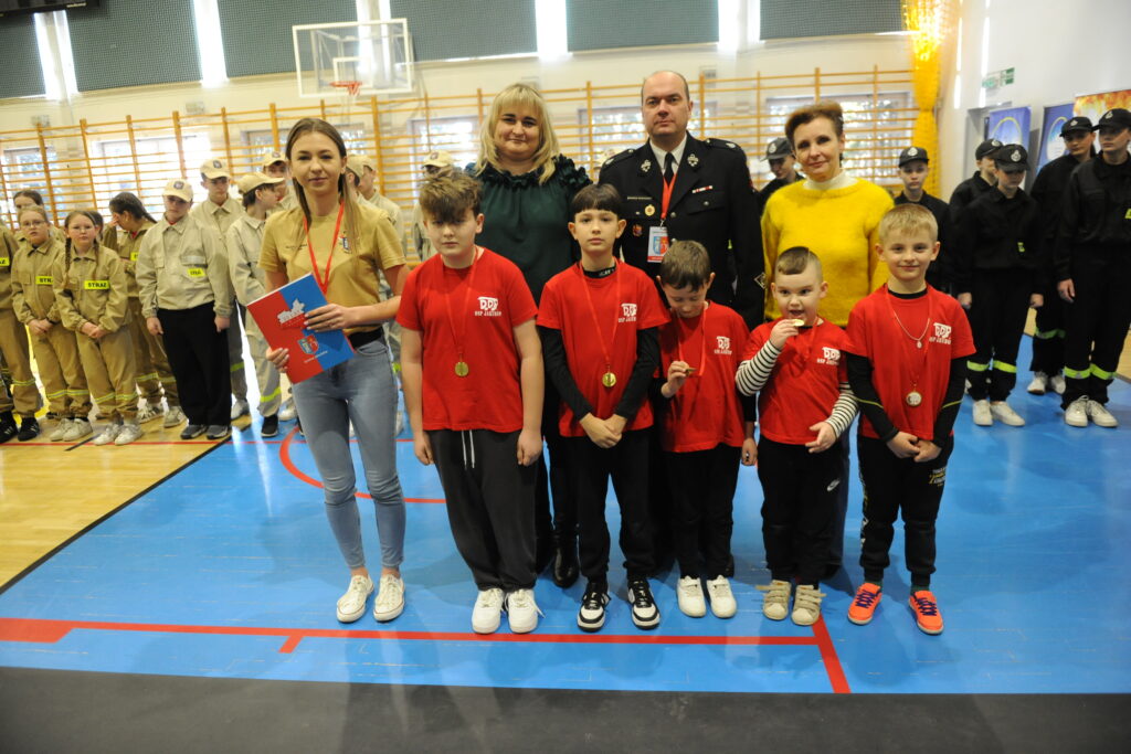 Grupa dzieci i dorosłych pozują na sali gimnastycznej. Mają czerwone medalami, a w tle stoją strażacy. Dominują kolory czerwony, niebieski i brązowy.