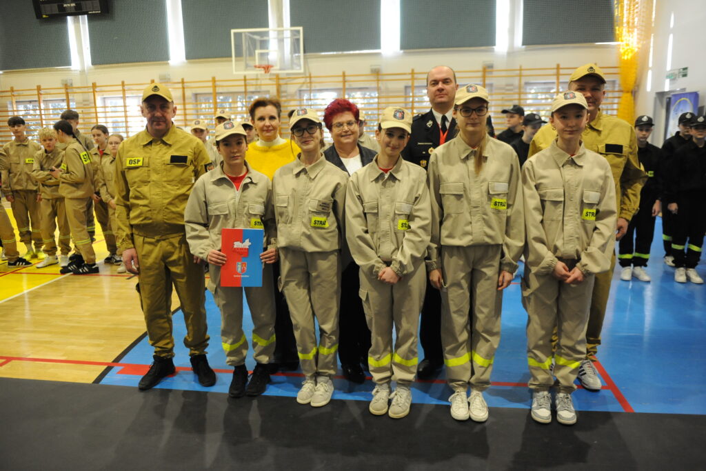 Grupa osób w mundurach strażackich stoi w sali gimnastycznej. Tło jest jasne z oknami i drewnianymi barierkami. Przeważają kolory beżowy i niebieski.