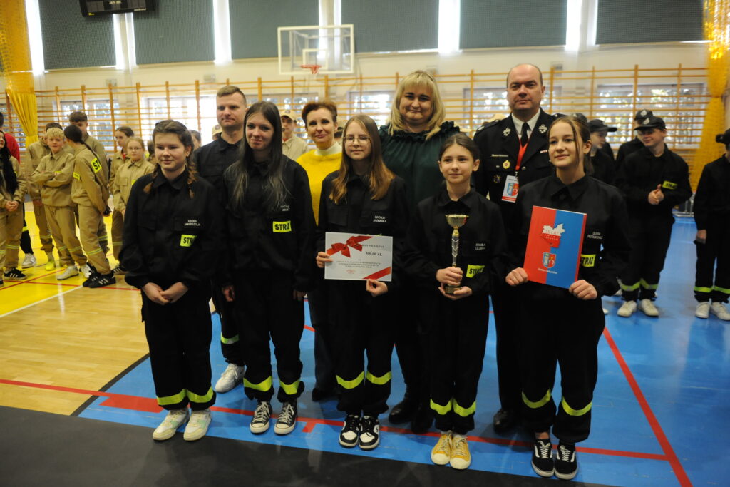 Grupa młodzieży w czarnych mundurach strażackich stoi na sali gimnastycznej. Środkowa osoba trzyma puchar i certyfikat. W tle widać osoby w beżowych mundurach oraz koszykówkę.