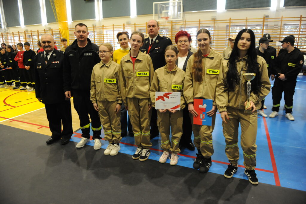 Grupa młodych strażaków w beżowych mundurach stoi na niebieskim tle hali sportowej. Trzymają dyplomy i puchar. W tle inni uczestnicy.