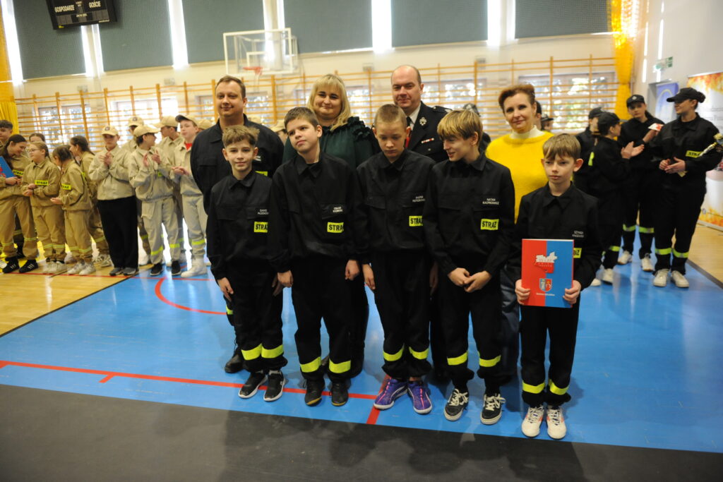 Grupa osób w mundurach strażackich stoi na niebieskim parkiecie sali gimnastycznej. Tło jest jasne z dużymi oknami. Po lewej stronie widać dzieci w beżowych uniformach.