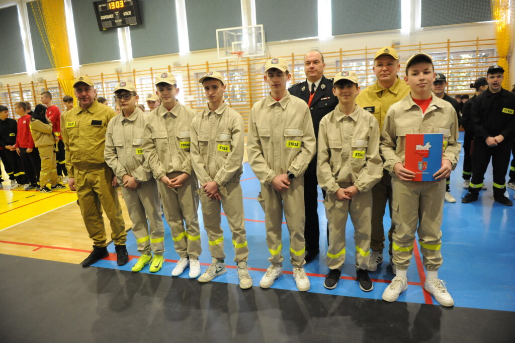 Grupa młodych strażaków w beżowych mundurach stoi na niebieskiej podłodze hali sportowej. Tło to jasne ściany z drewnianymi drabinkami. Jeden z uczestników trzyma czerwononiebieską teczkę.