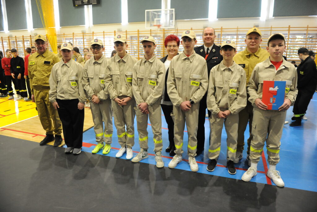 Grupa młodych ludzi w beżowych mundurach stoi w rzędzie. Tło to sala gimnastyczna z widoczną podłogą w jasnych kolorach. Osoba z folderem o czerwono-niebieskiej okładce po prawej.