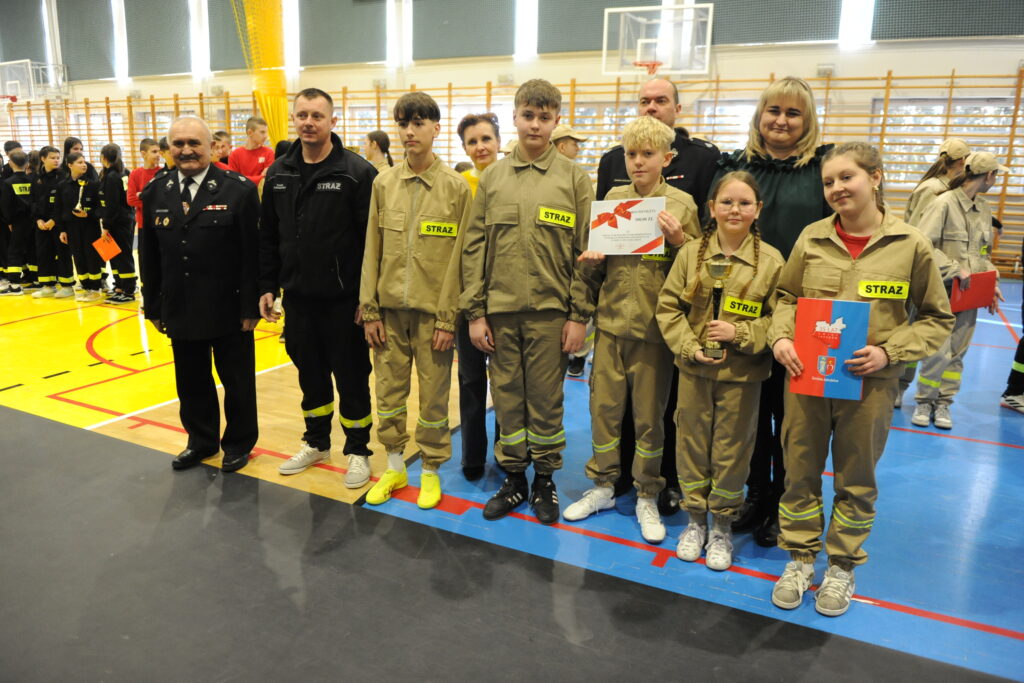 Grupa osób w strojach strażackich stoi na hali sportowej. Tło jest jasne, a podłoga kolorowa. Kilku uczestników trzyma nagrody.