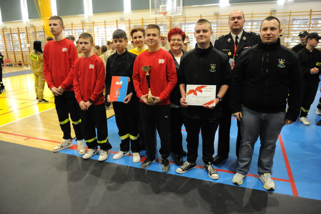 Grupa młodzieży i dorosłych w czerwonych i czarnych strojach stoi w sali gimnastycznej. Jeden z nich trzyma puchar, inny dyplom. W tle widoczne są jasne ściany i drewniane drabinki.