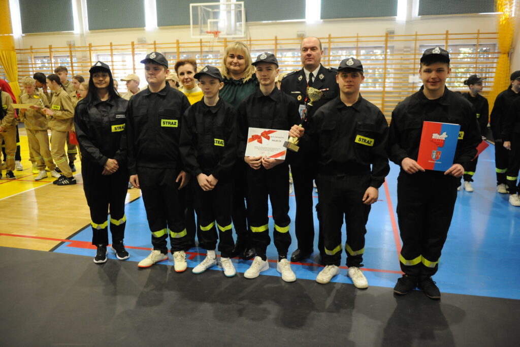 Grupa młodych strażaków w czarnych mundurach z trofeum na tle sali gimnastycznej. W tle stoją osoby w beżowych uniformach, ściany są jasne. Na podłodze widać niebieskie i żółte oznaczenia.