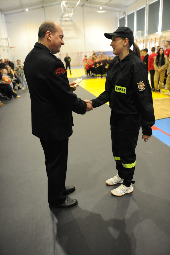 Dwaj strażacy w mundurach uściskują dłonie na hali sportowej. W tle grupa ludzi, jasne ściany, podłoga żółto-niebieska.
