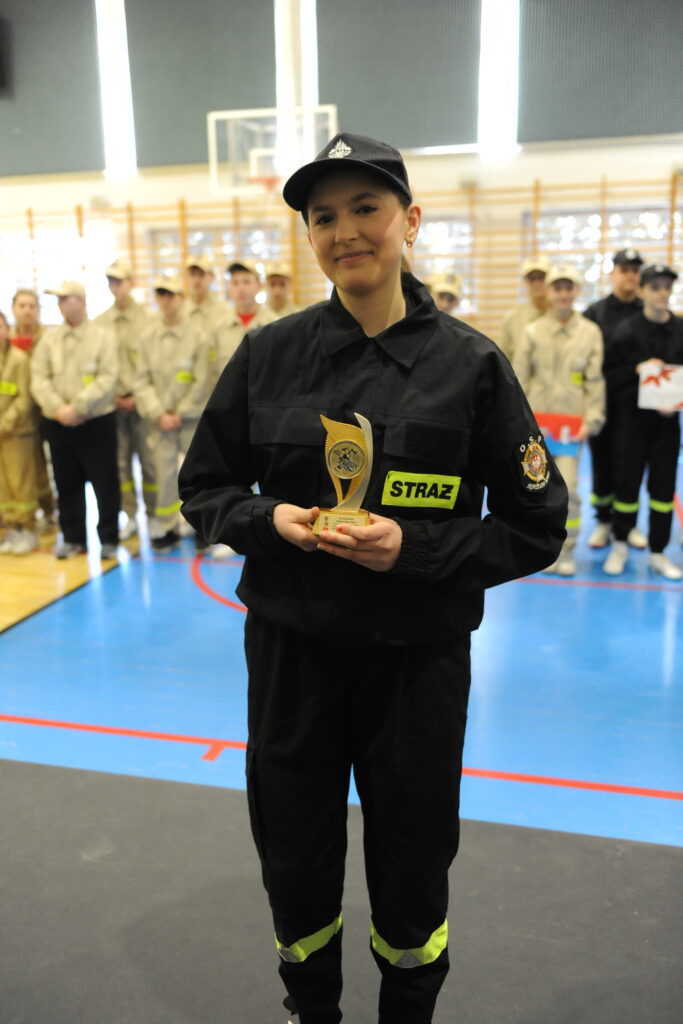 Strażak w czarnym mundurze trzyma trofeum. W tle grupa w beżowych mundurach. Podłoga niebiesko-czarna w sali gimnastycznej.