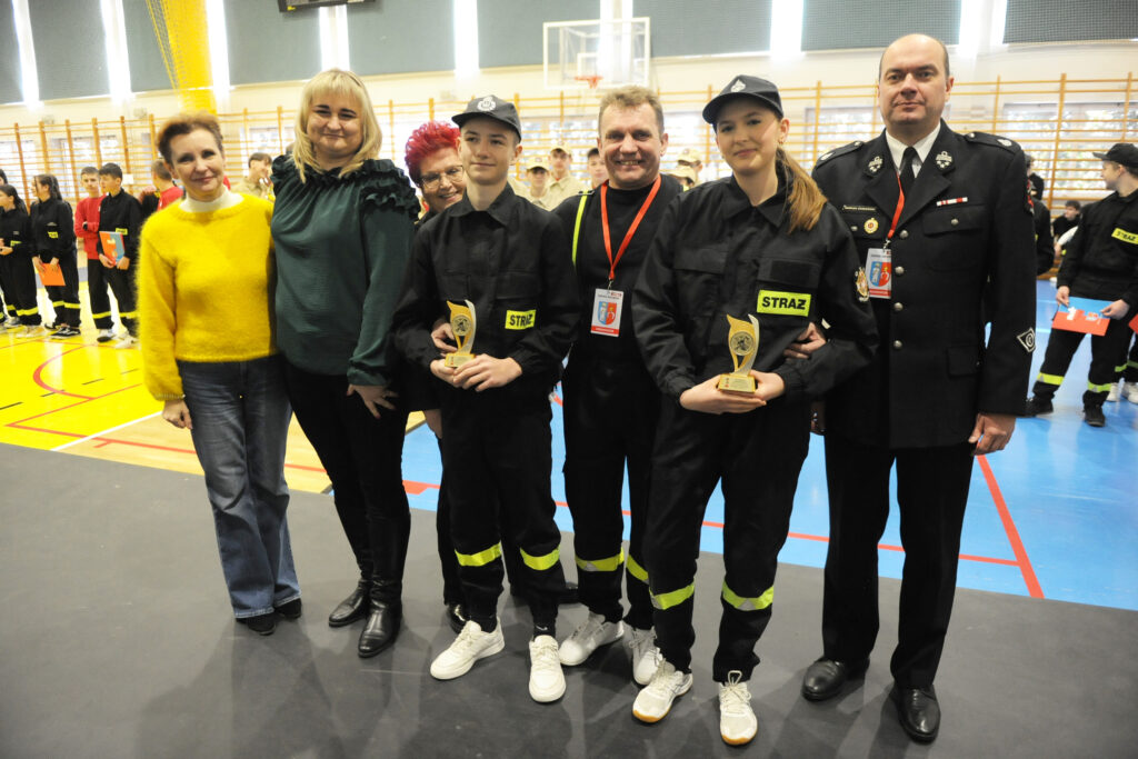 Grupa osób w strojach strażackich trzyma nagrody, stoją na boisku sportowym. Tło jasne, dominuje żółty i niebieski. W tle kolejny rząd osób.